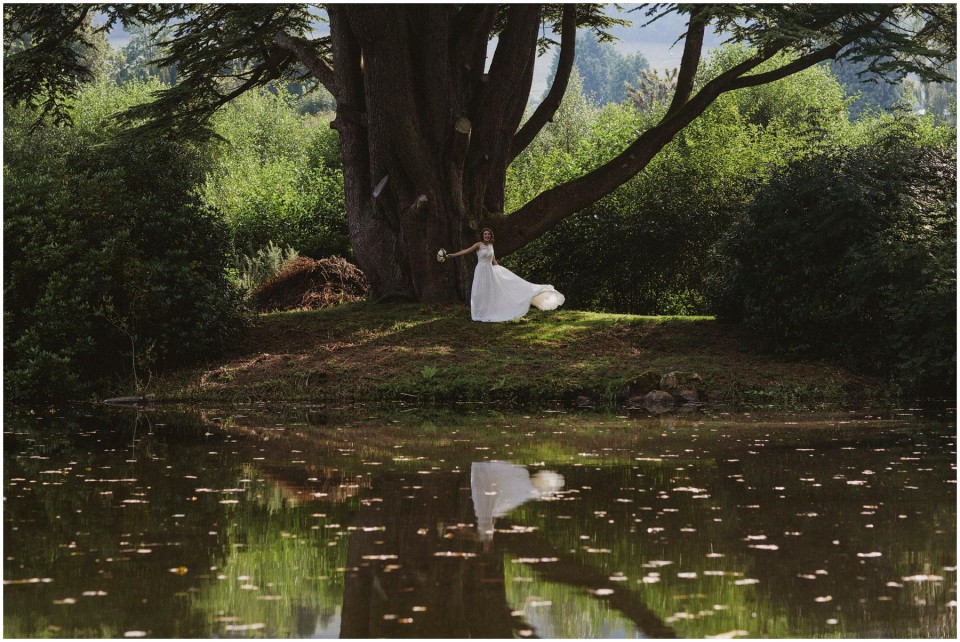 Bride in flowing white dress beneath towering tree, reflected in still pond, documentary wedding photography.