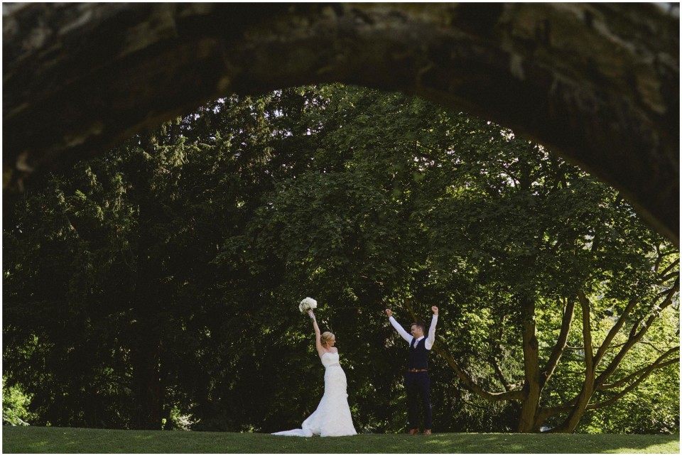 Bride and groom raise arms beneath sweeping trees, bouquet lifted high