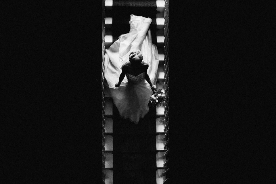 Bride descending staircase, dress flowing, bouquet in hand, captured in quiet black-and-white wedding photography.