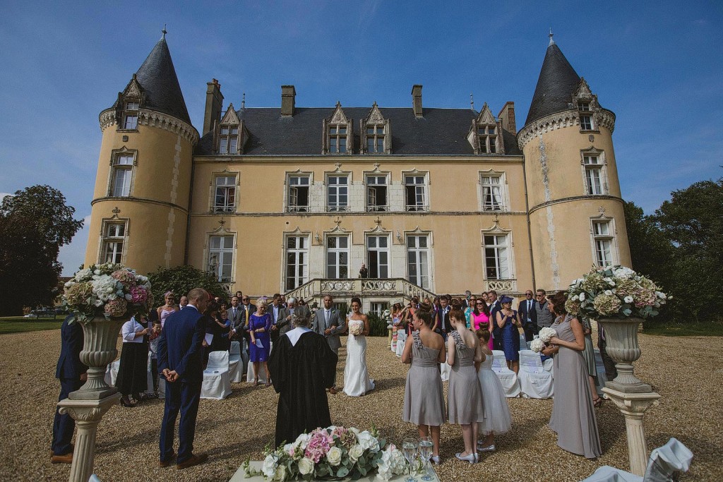 Guests gather for an outdoor wedding ceremony before a sunlit French château.
