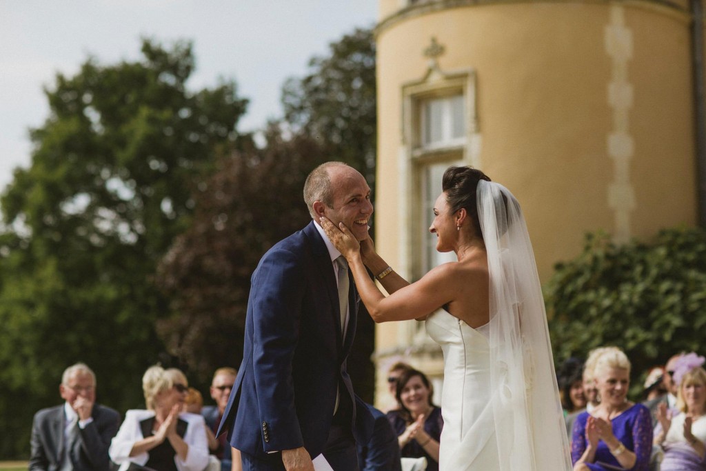 Bride cups groom’s face, both laughing as guests applaud in sunlit garden wedding ceremony.