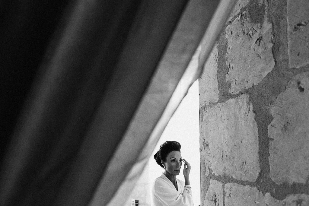 Bride applies mascara beside a stone wall, morning light falling softly – documentary wedding photography.