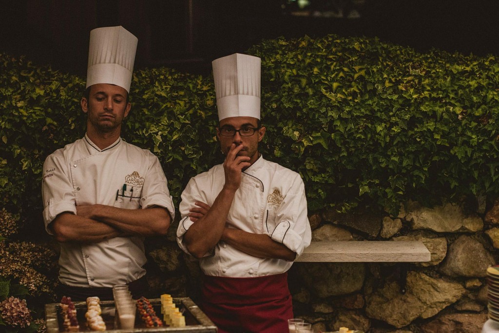 Two chefs in tall white hats stand quietly behind dessert table at evening wedding reception.