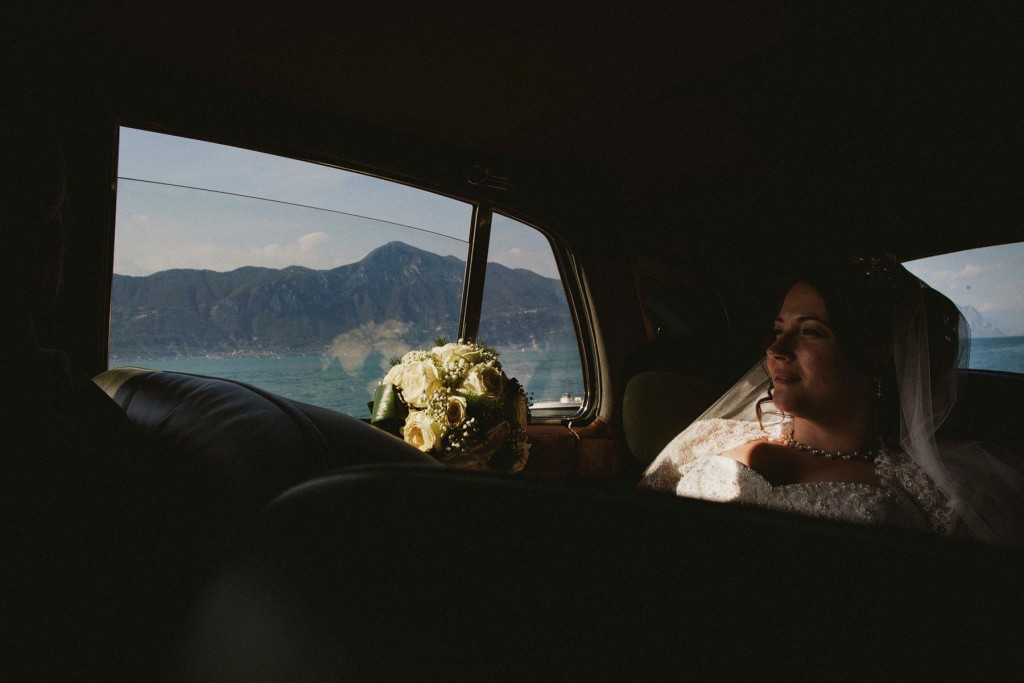 Bride gazes through car window at lake and mountains, bouquet resting beside her — documentary wedding photography.