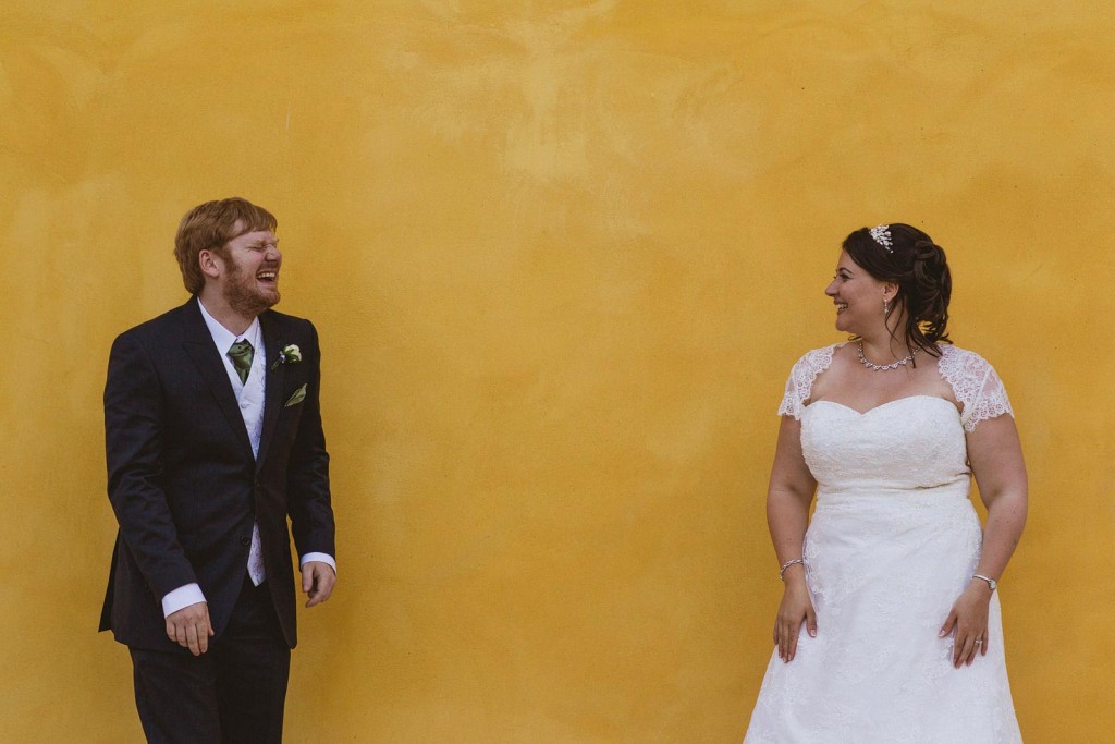 Bride and groom laughing across a mustard wall on their wedding day.