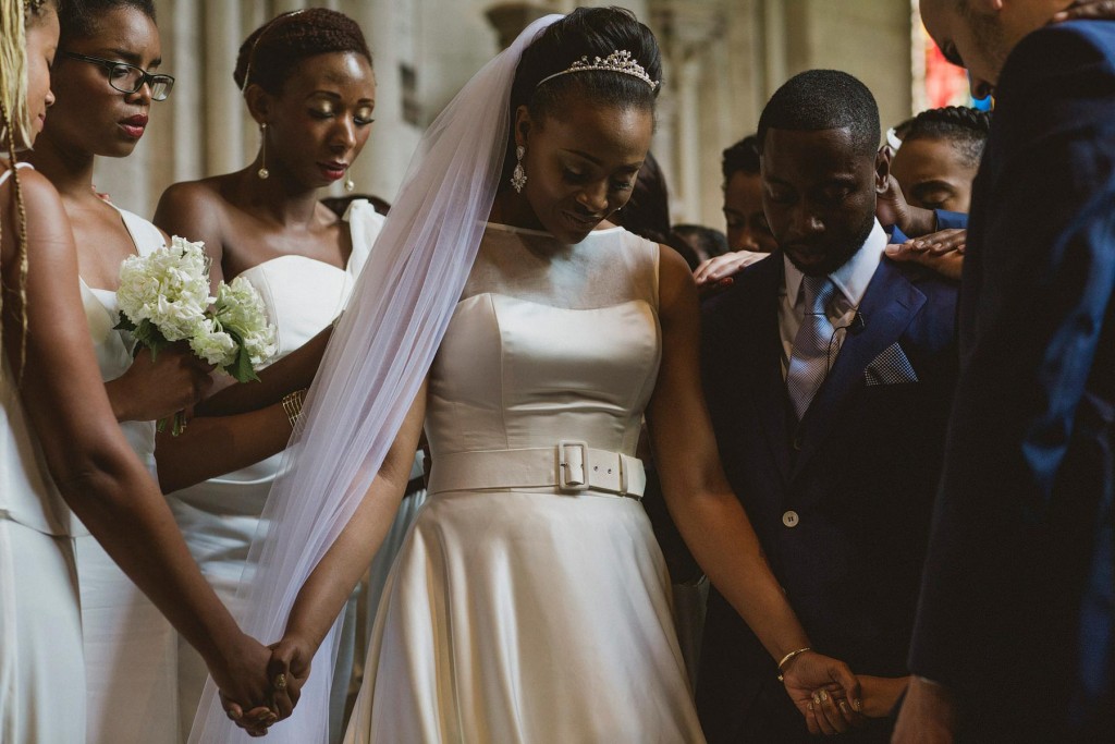 Bride and groom hold hands with family, heads bowed during church wedding prayer.