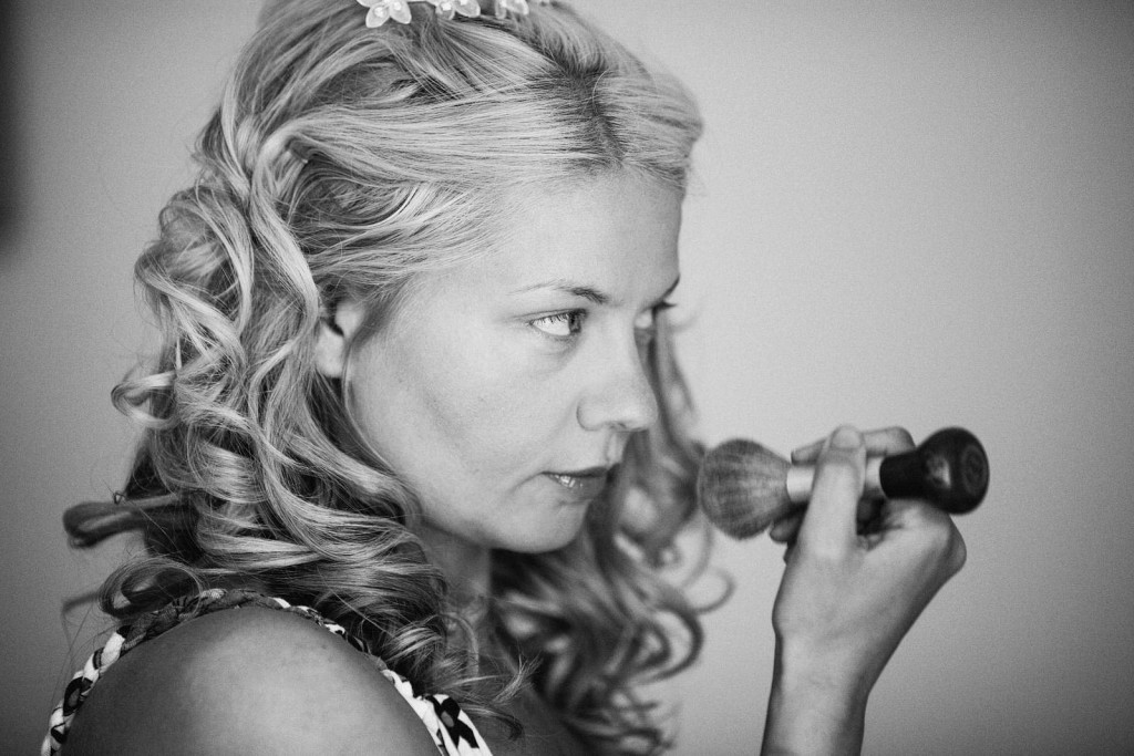 Bride applying blusher, softly lit, curls pinned back, quiet anticipation before ceremony in wedding photography.