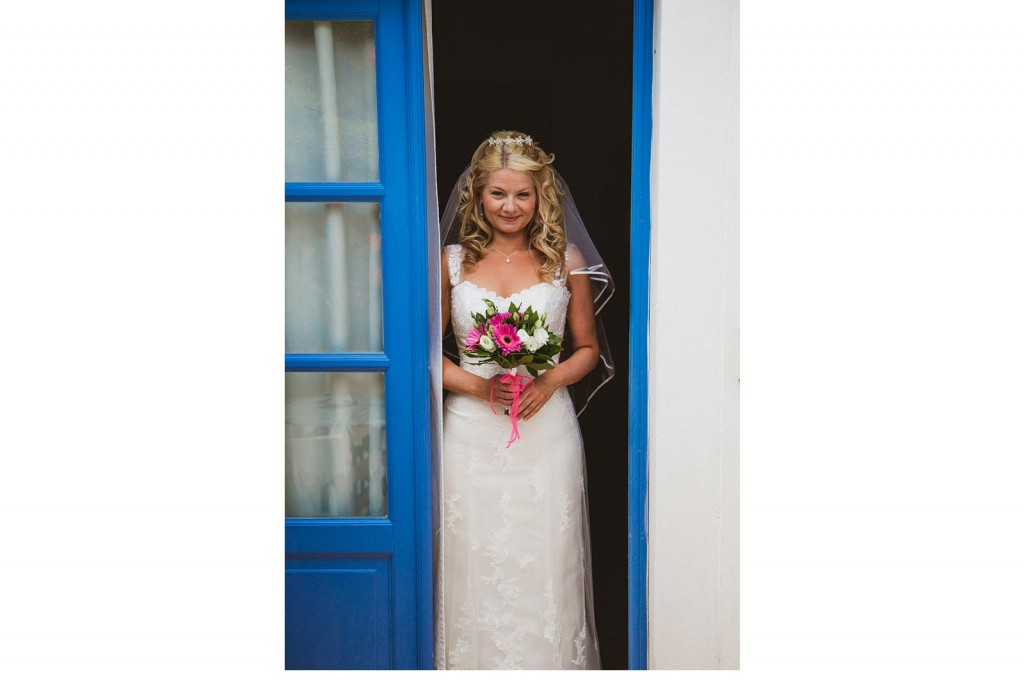 Bride in lace gown stands in blue doorway, holding bright bouquet, soft smile documentary wedding photography moment.