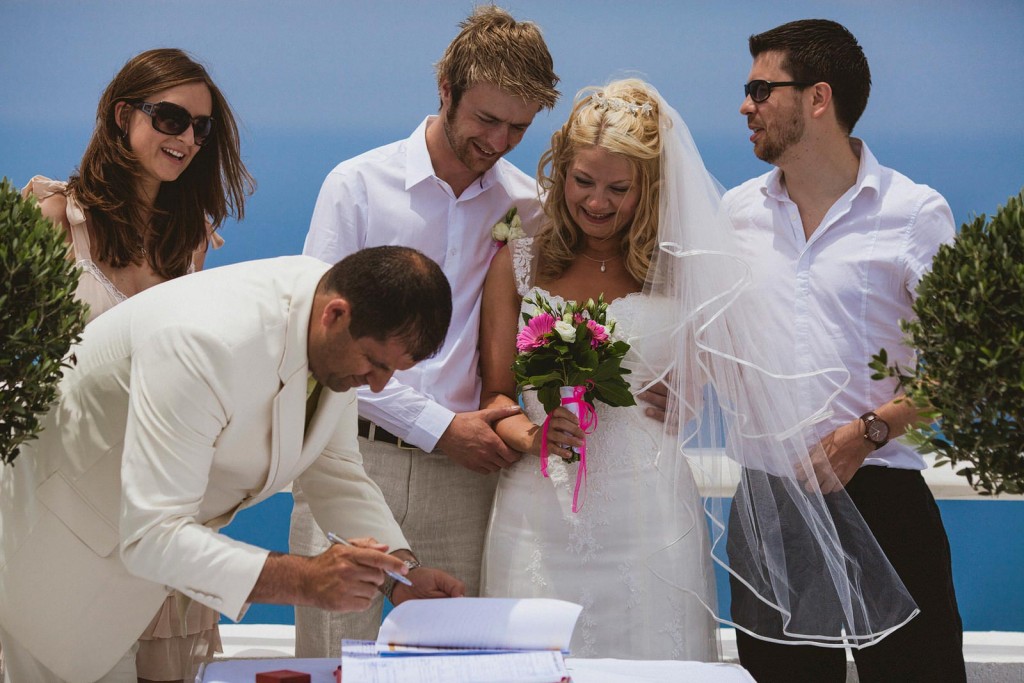 Groom signs wedding register as bride smiles with bouquet, friends gathered beneath bright blue sky.