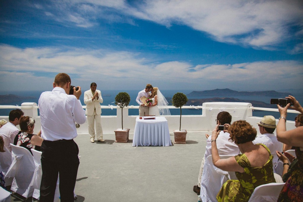 Bride and groom kiss during cliffside wedding ceremony as guests raise cameras beneath wide blue sky.