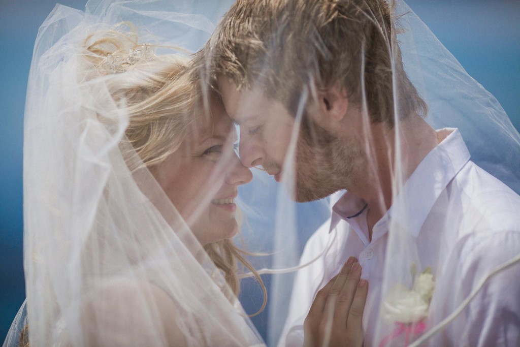 Bride and groom rest foreheads together beneath a veil, smiling softly in sea breeze wedding photography.