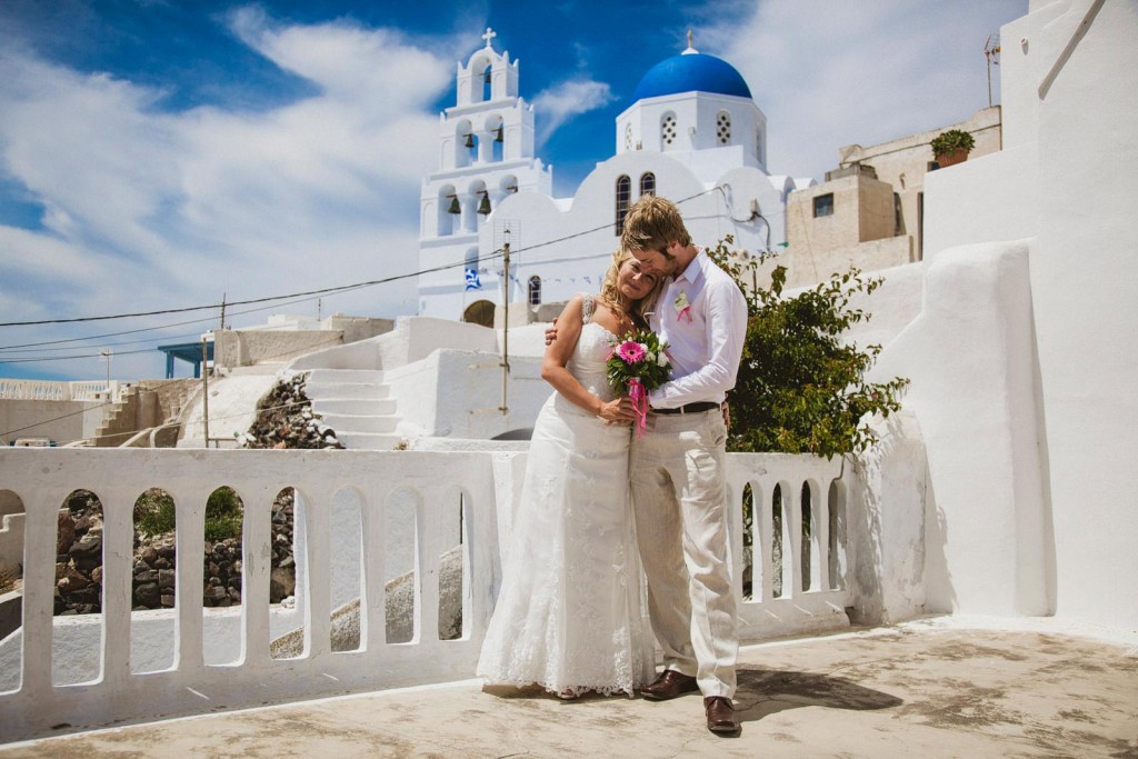 Bride and groom embrace on sunlit Santorini terrace, white church and blue dome behind.