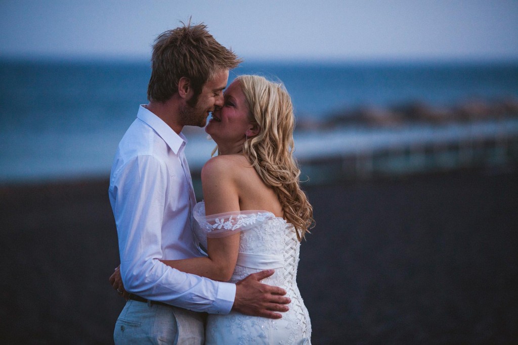 Bride and groom share a quiet kiss at dusk by the sea, laughter between them in documentary wedding photography.