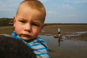 Toddler near camera while another child stands on a rock in shallow beach water.