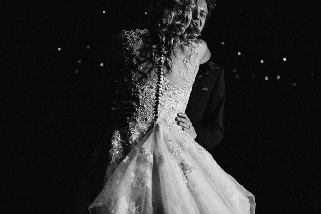 Bride and groom embrace during evening wedding dance, her beaded gown catching soft light.