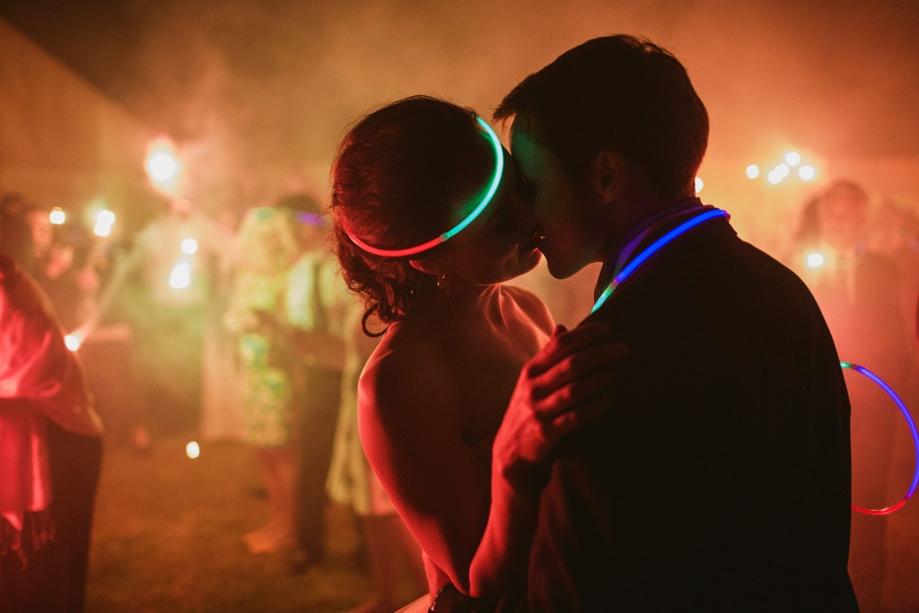 Couple kissing on dance floor, glow necklaces shining at night wedding reception.