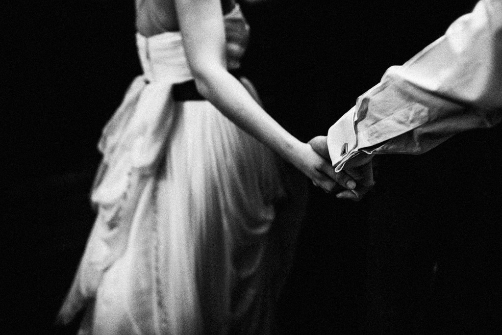 Black and white wedding photography of couple holding hands, bride’s gown softly gathered in shadow.