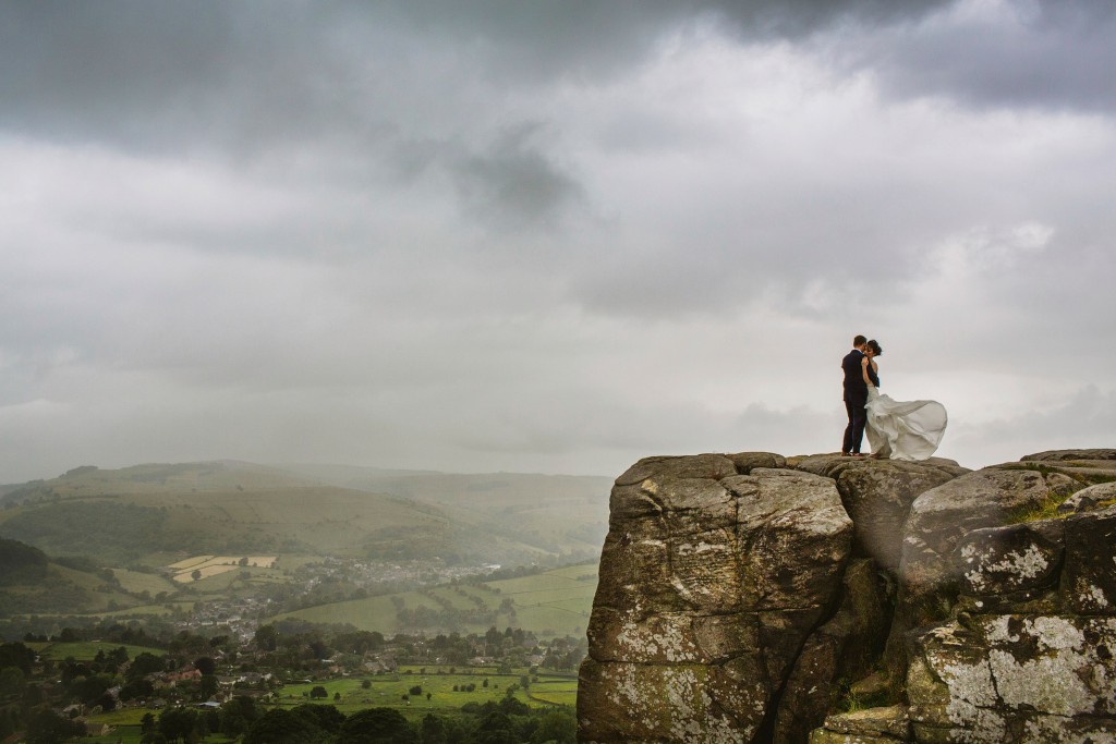 Bride and groom stand on rocky cliff, dress billowing above misty valley, documentary wedding photography.