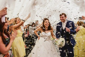 Bride and groom laughing beneath falling confetti as guests cheer around them at wedding celebration.