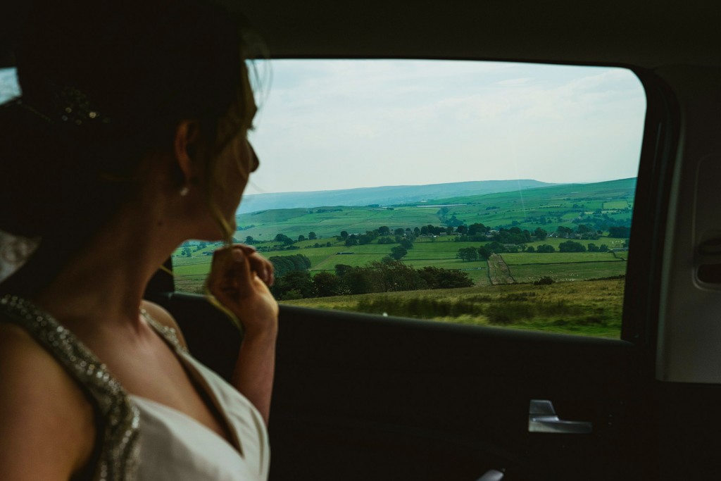 Bride gazes through car window at rolling green countryside, quiet moment during wedding day.