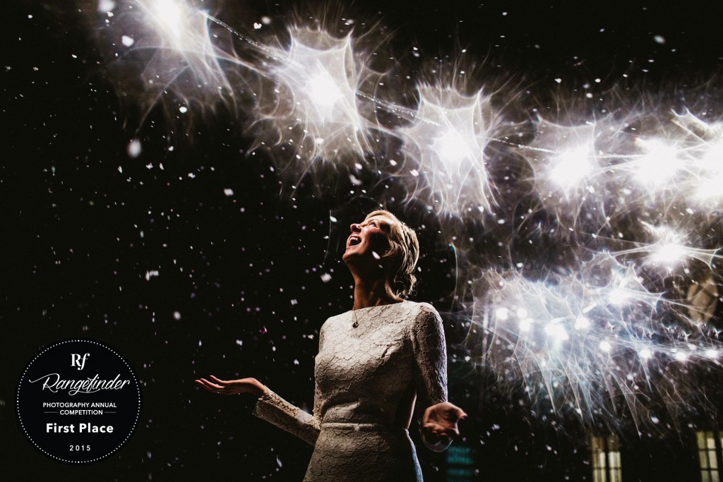Bride laughing beneath bright fireworks, confetti drifting through night sky during wedding celebration.