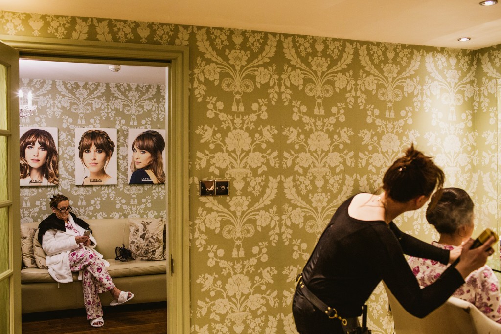 Bride's mother having hair styled while another woman waits on sofa, quiet wedding morning.