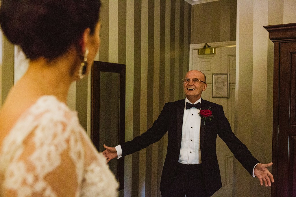 Father in tuxedo opens his arms, smiling at bride in doorway, wedding photography moment.