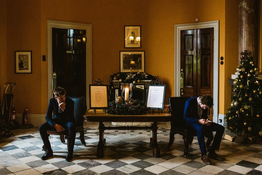 Two suited men sit apart in a candlelit hall beside a decorated Christmas tree, checking phones.