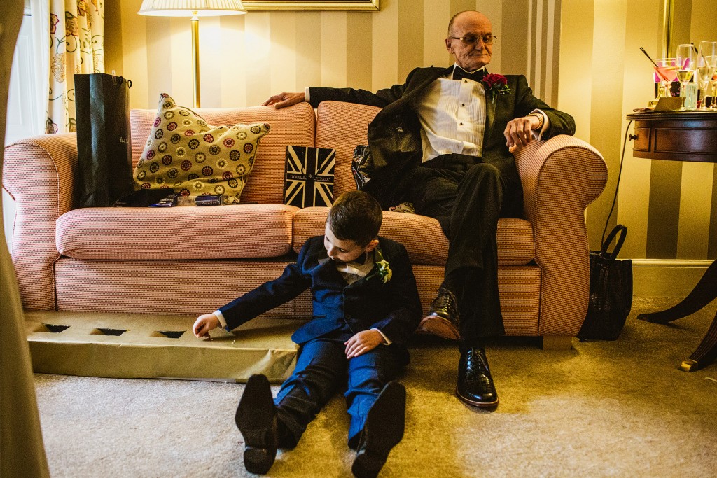 Young boy in suit opens a gift on carpet as older man rests on sofa, wedding morning.