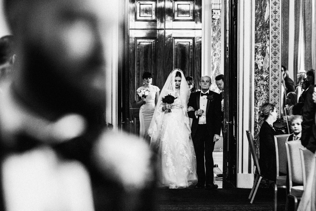 Bride enters wedding ceremony with her father, guests watching quietly in a grand hall.