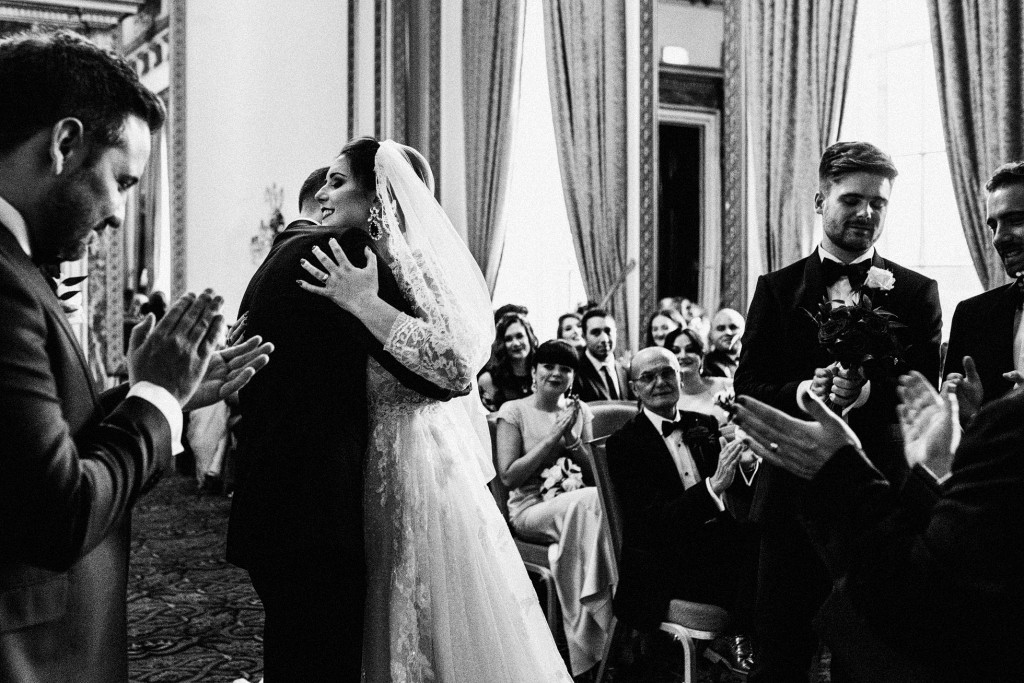 Bride and groom embrace as guests applaud during indoor wedding ceremony.