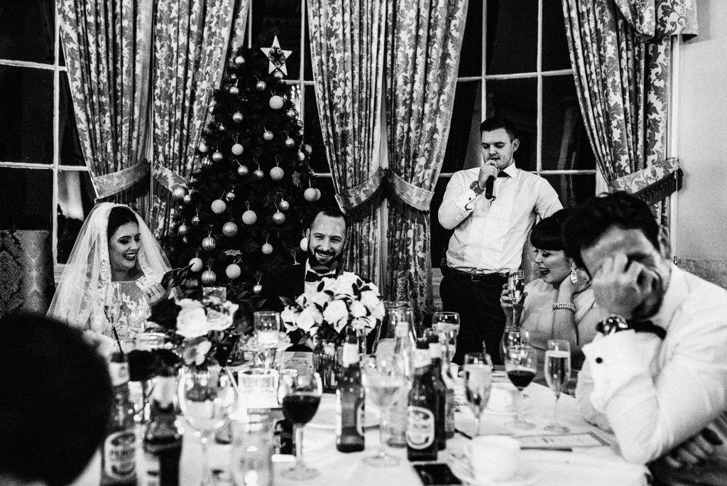 Bride and guests laughing during heartfelt speech at candlelit wedding reception beside Christmas tree.