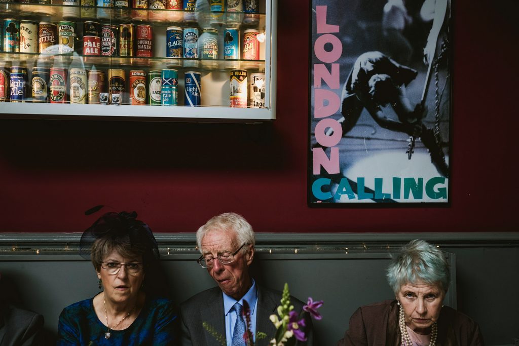 Three wedding guests sit quietly beneath vintage beer cans and a bold poster, lost in thought.