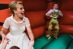 Bride laughing inside bouncy castle as child in dinosaur costume jumps nearby at wedding.