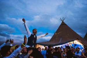 Guests lift groom high, arms wide, cheering beneath twilight tipi at wedding reception.