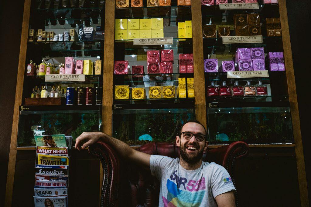 Man laughing in red leather chair beneath shelves of colourful grooming products and magazines.