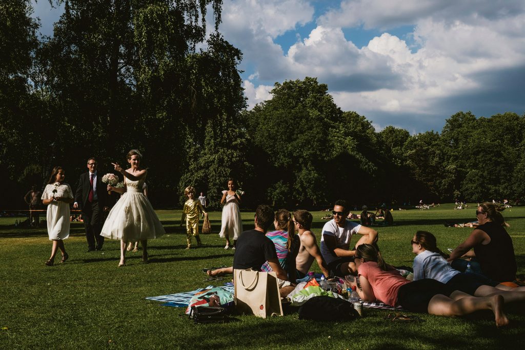 Bride and groom walk across sunny park lawn toward friends picnicking on blankets, documentary wedding photography moment.