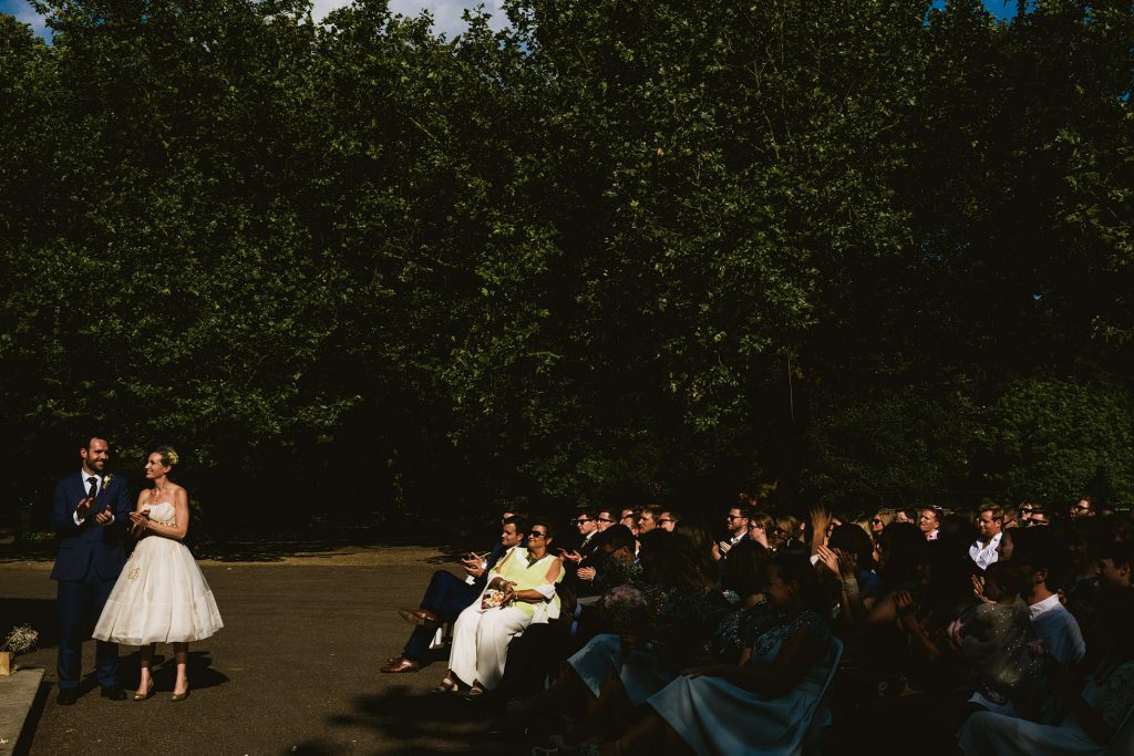 Bride and groom standing in sunlight as guests applaud beneath tall green trees, documentary wedding photography.