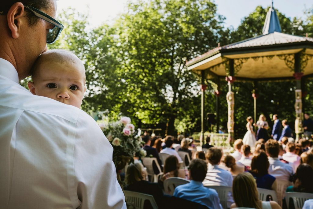 Baby resting on man's shoulder as outdoor wedding ceremony unfolds beneath leafy trees.