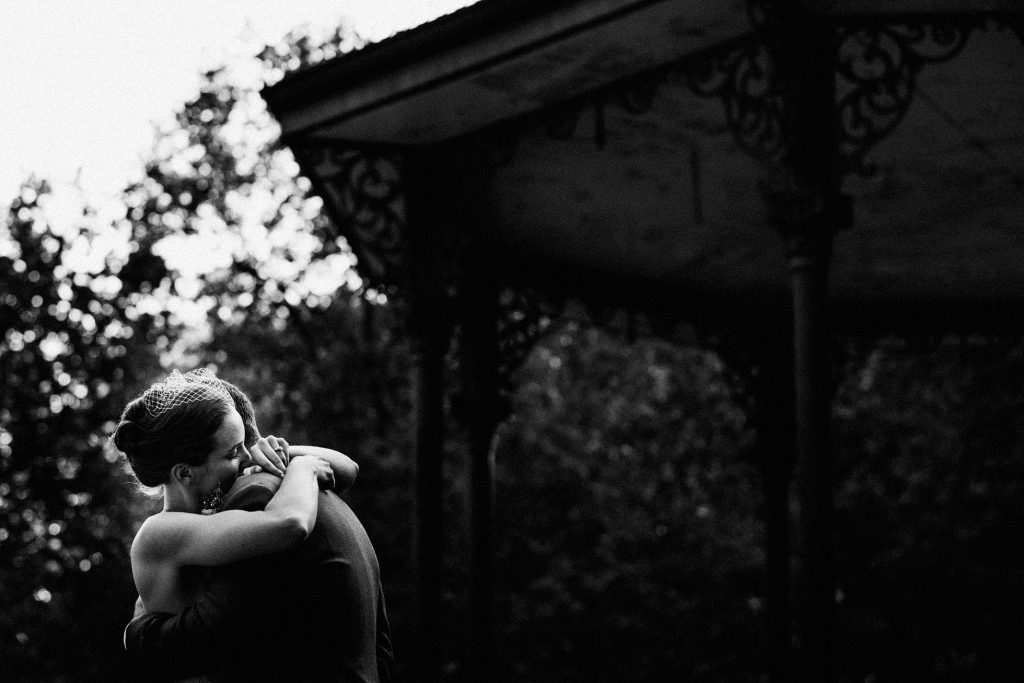 Bride in veil embraces partner beneath ornate pavilion at dusk, quiet black and white wedding photography moment.