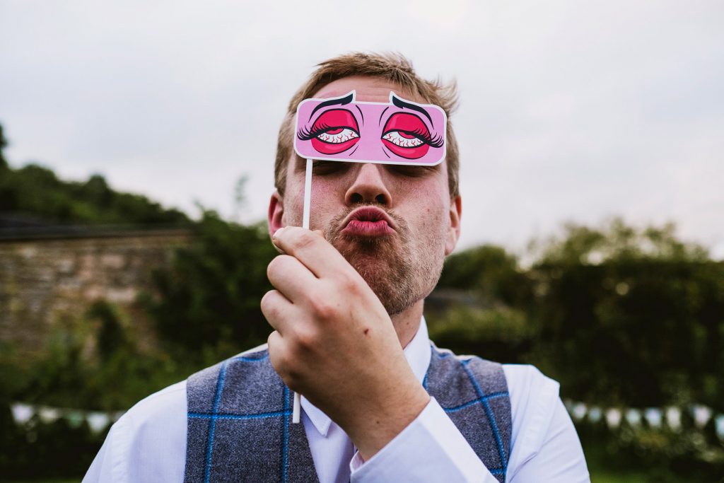 Guest holds cartoon eye mask over his face, puckering lips in garden during wedding celebration.