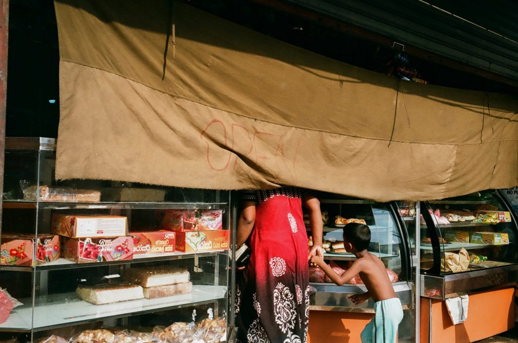 Woman in red dress buys bread as child holds her hand beneath shop awning.