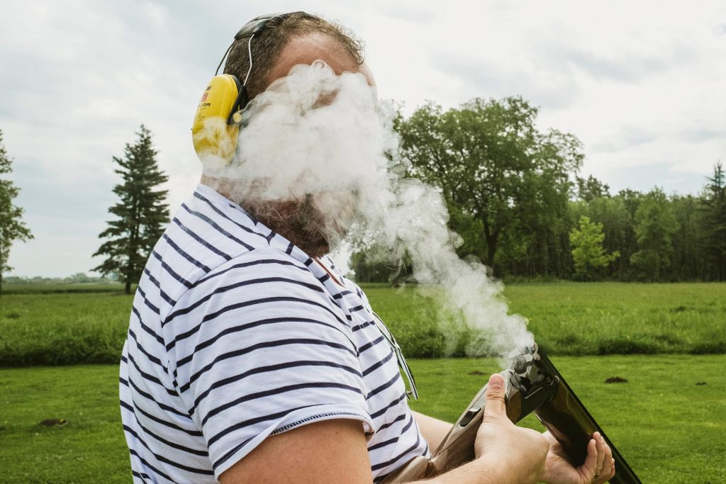 Man in striped shirt and ear defenders holds shotgun as smoke drifts across his face in open field.