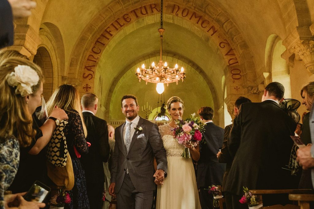 Bride and groom walk down church aisle smiling, guests gathered close, warm light glowing overhead during wedding ceremony.