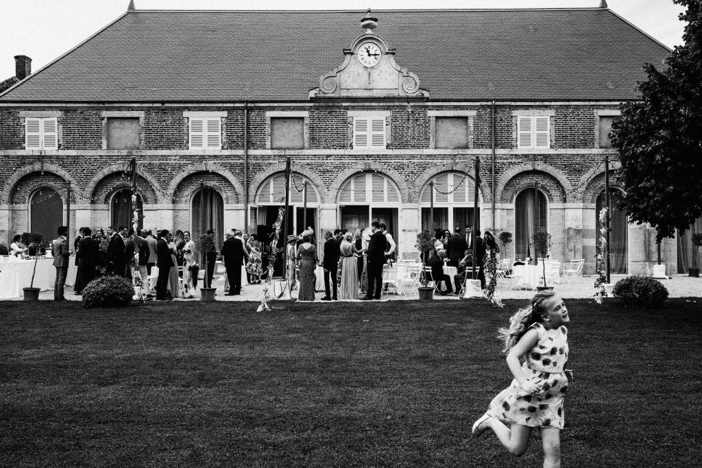 Young girl runs across lawn as guests mingle outside grand brick building during wedding reception.