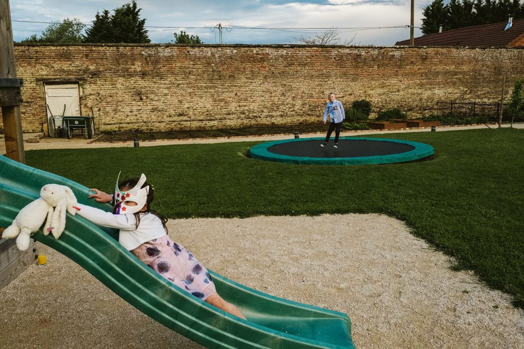 Child in rabbit mask slides down garden slide while another bounces on trampoline.
