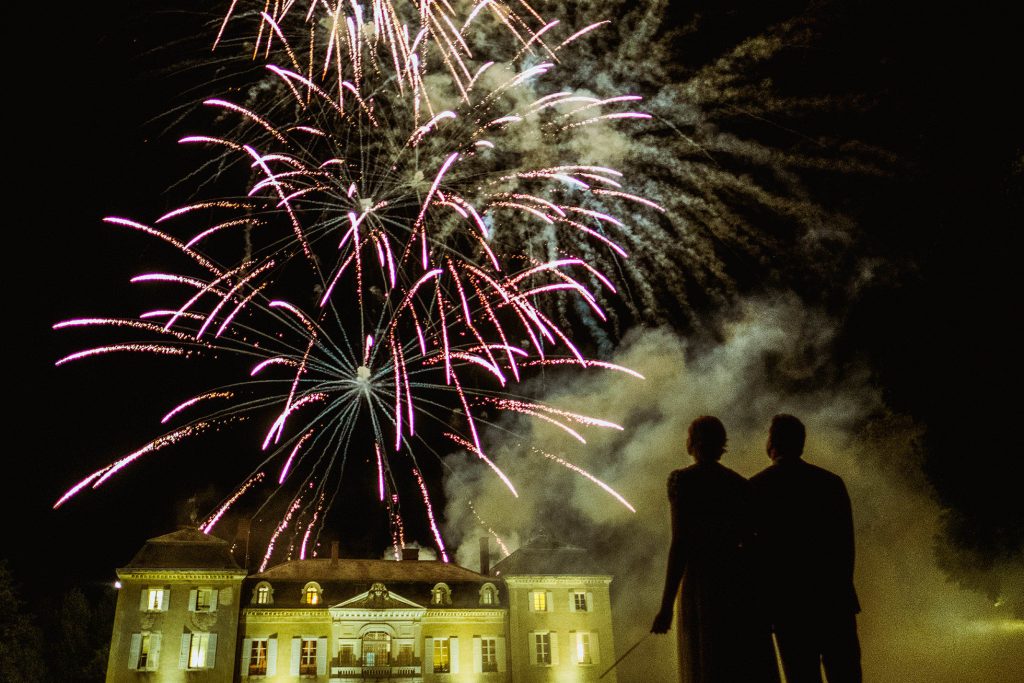 Silhouetted couple watch fireworks above grand country house at night, smoke drifting through wedding celebration.