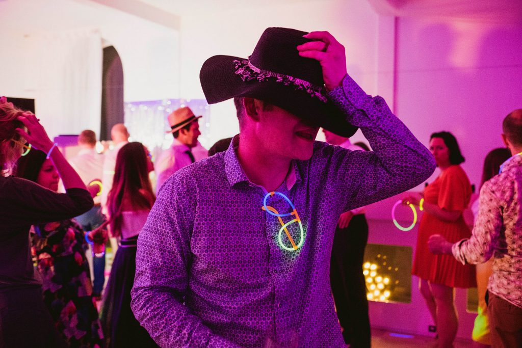 Guest in cowboy hat dancing under pink lights at wedding reception, glow sticks bright against his shirt.