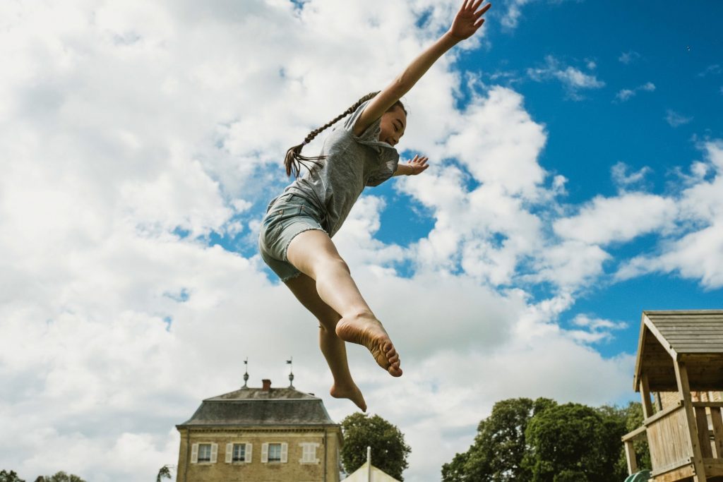 Barefoot child leaps into bright blue sky above garden house, arms wide in carefree joy.