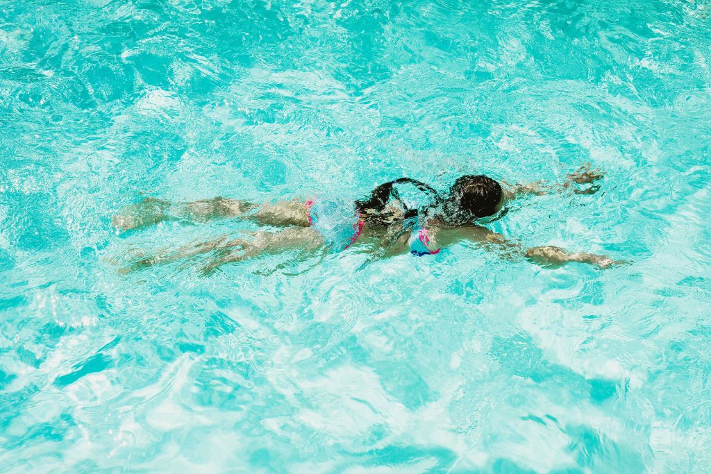 Woman floating face down in bright turquoise swimming pool, arms outstretched, sunlight rippling across water.